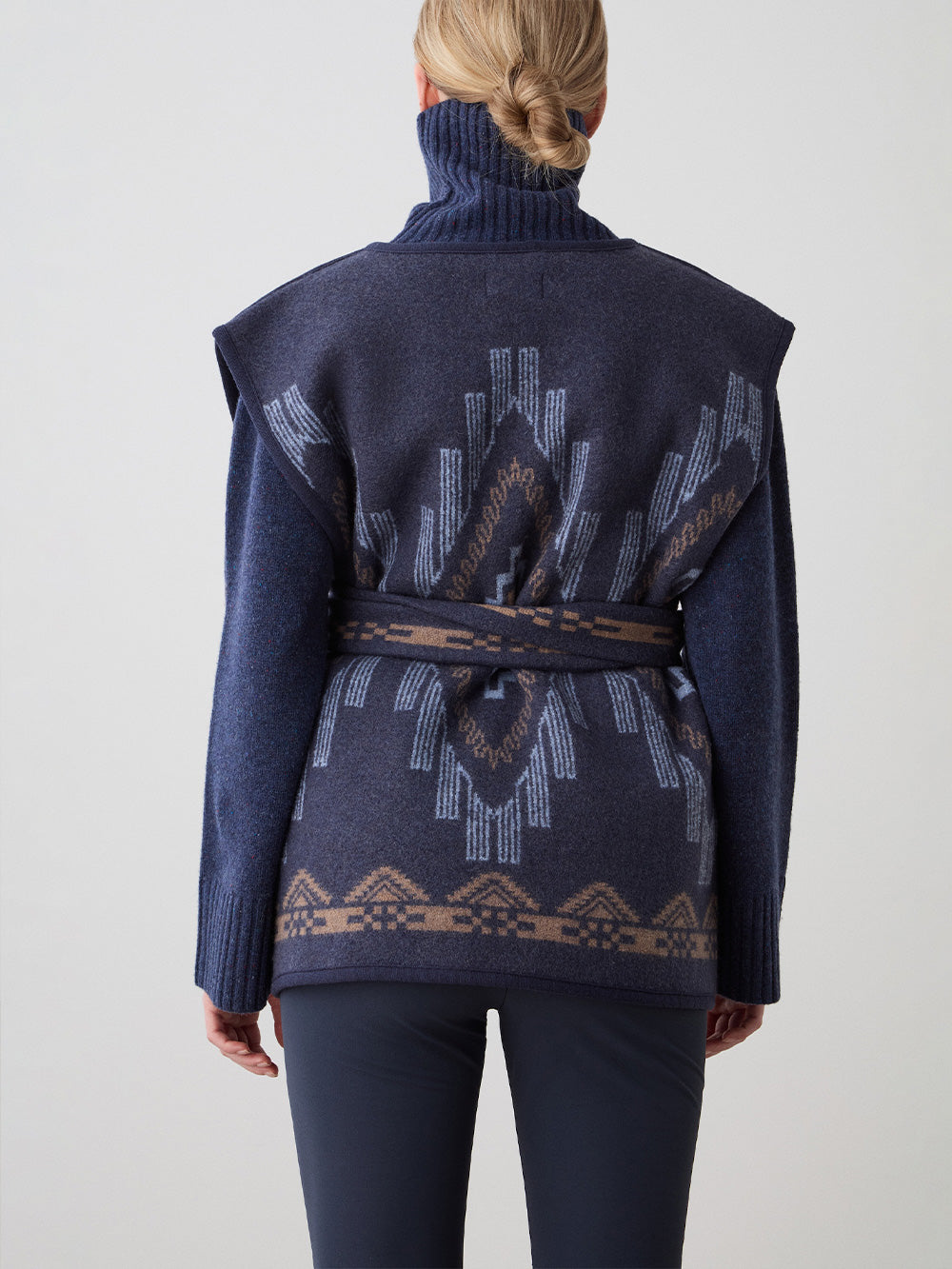 We Norwegians_FW25_Womens | Model is wearing the Pioneer vest | Navy blue in color with a jacquard pattern | Vest has a matching belt to cinch the waist | Back view on a white background