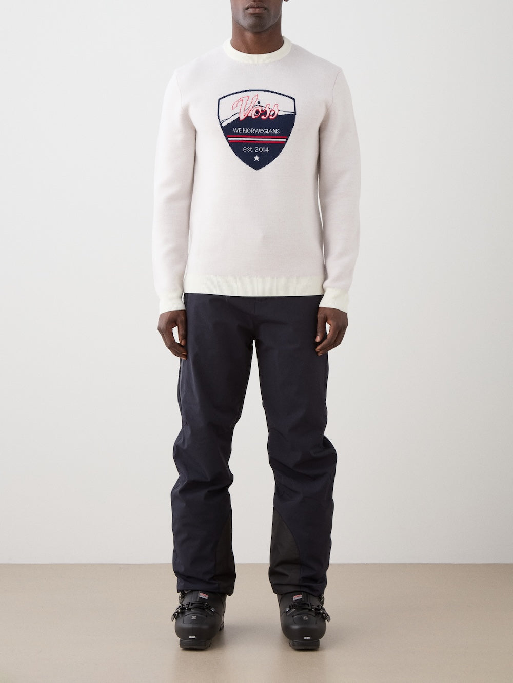We Norwegians | FW25 | Mens Afterski Crewneck_Cream | Model wearing a knitted sweater with a design and logo at the front with black ski pants | Shot against an off-white background | Front view